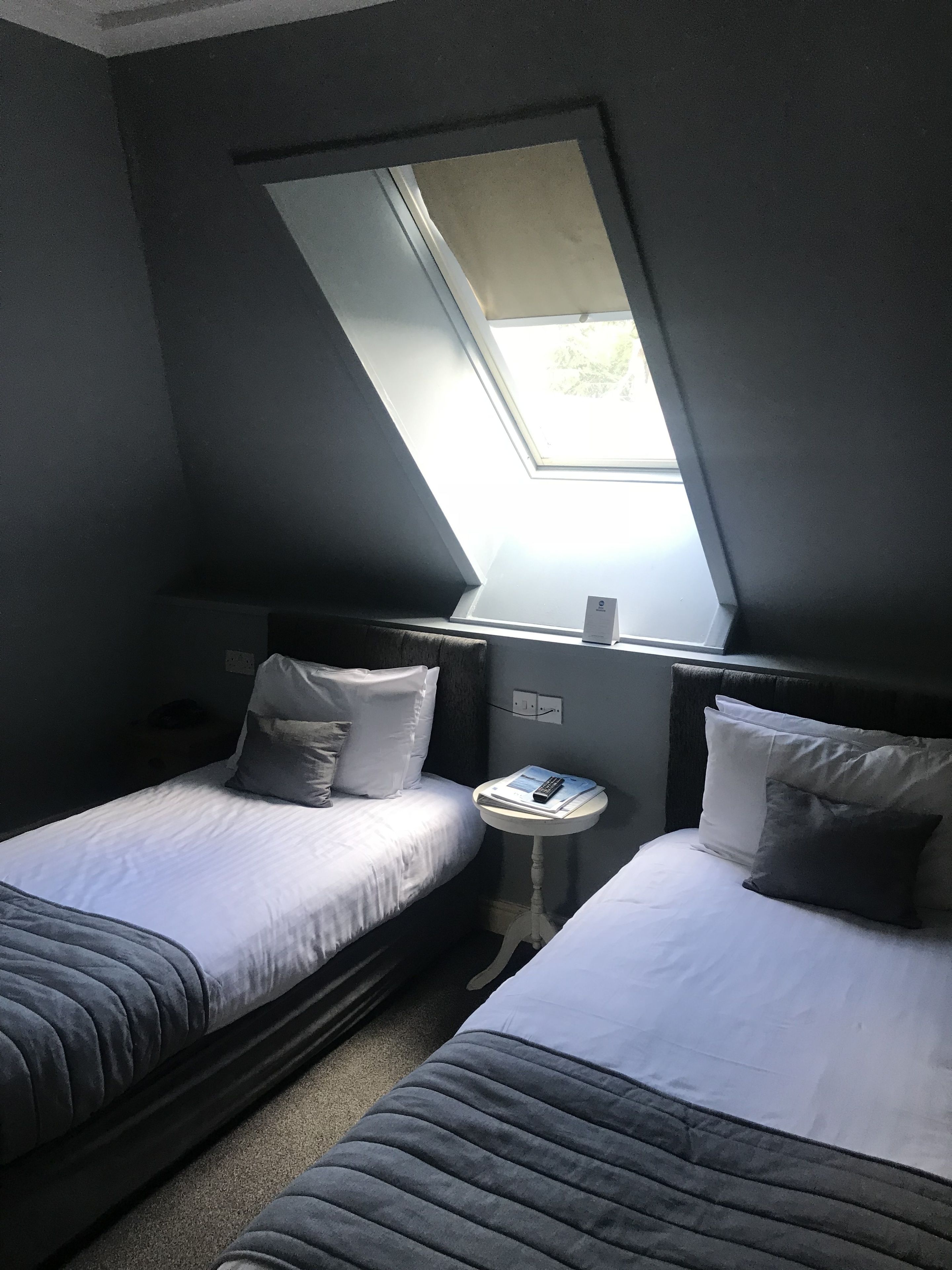 Une petite pièce peu éclairée, avec deux lits simples, une petite table et une lucarne.