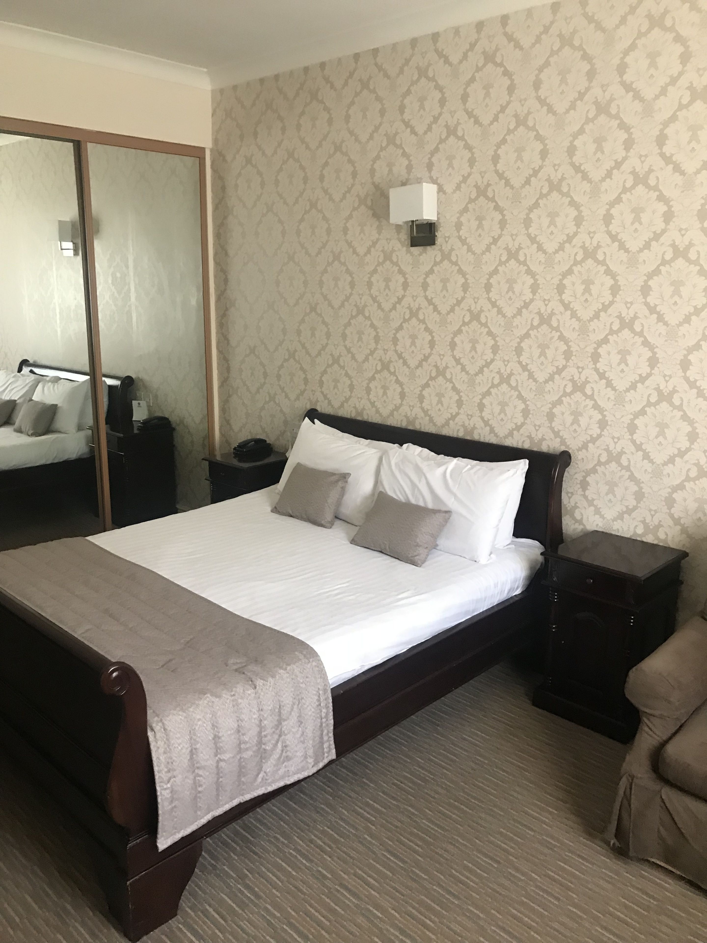 Una cama bien hecha con edredón blanco y dos almohadas, cabecera de madera, mesita de noche con lámpara y un espejo en la pared.
