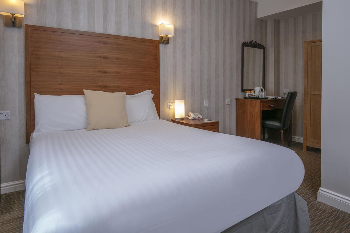 Hotellihuone, jossa on suuri sänky, puinen pääty, työpöytä tuolilla, peili ja yöpöytä lampulla.