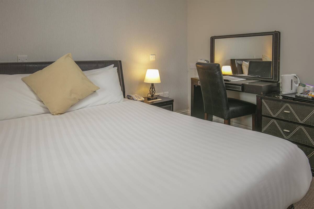 Hotellihuone, jossa on suuri sänky, työpöytä tuolilla, lamppu, peili ja lipasto.