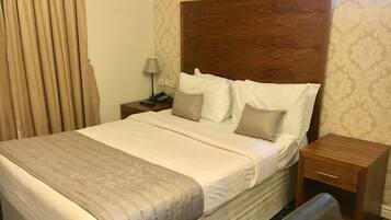 A hotel room with a large bed, a wooden headboard, a nightstand with a lamp, and a window with curtains.