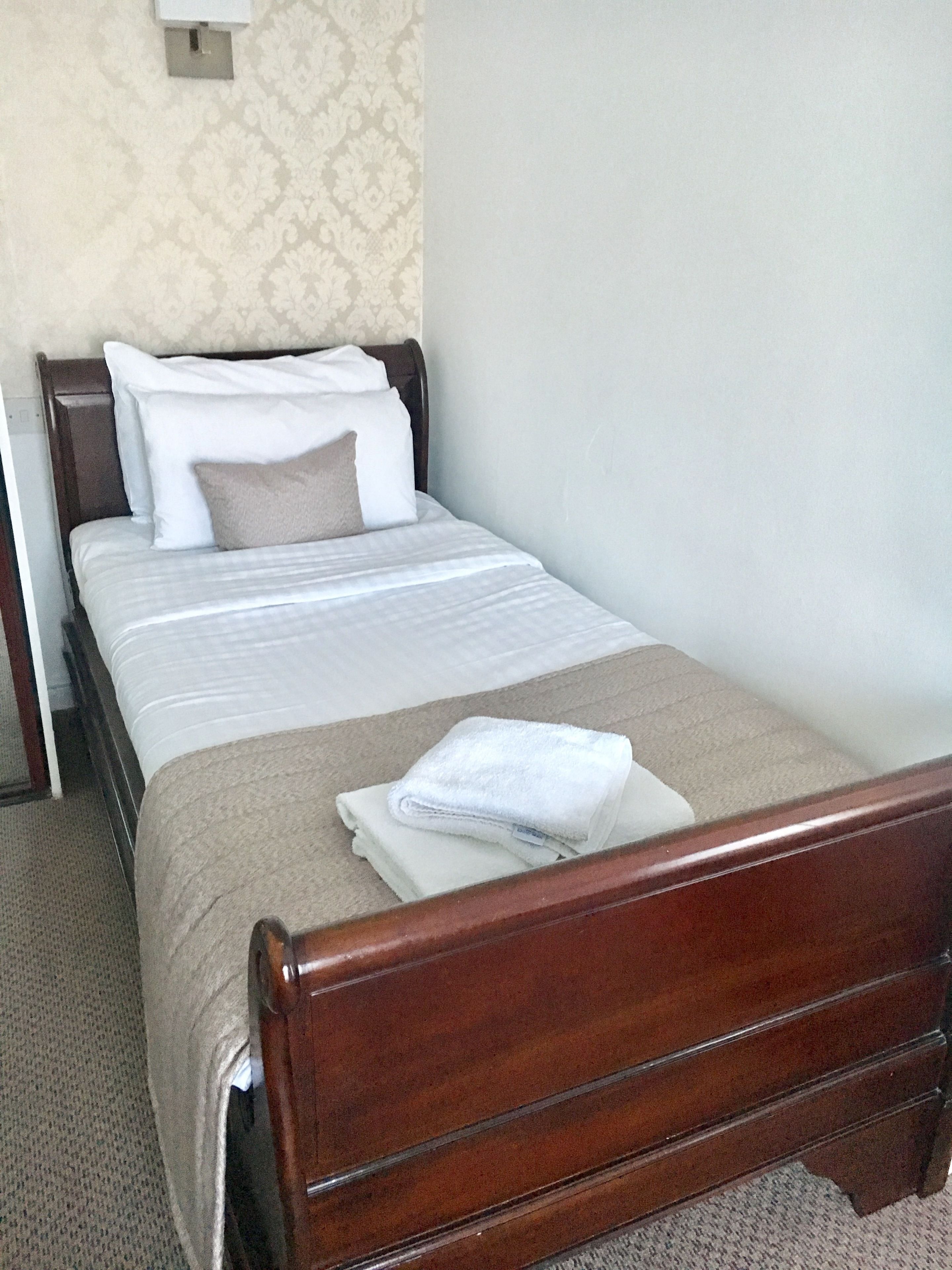 A neatly made bed with white and beige bedding, a wooden headboard, and a folded white towel on the footboard.