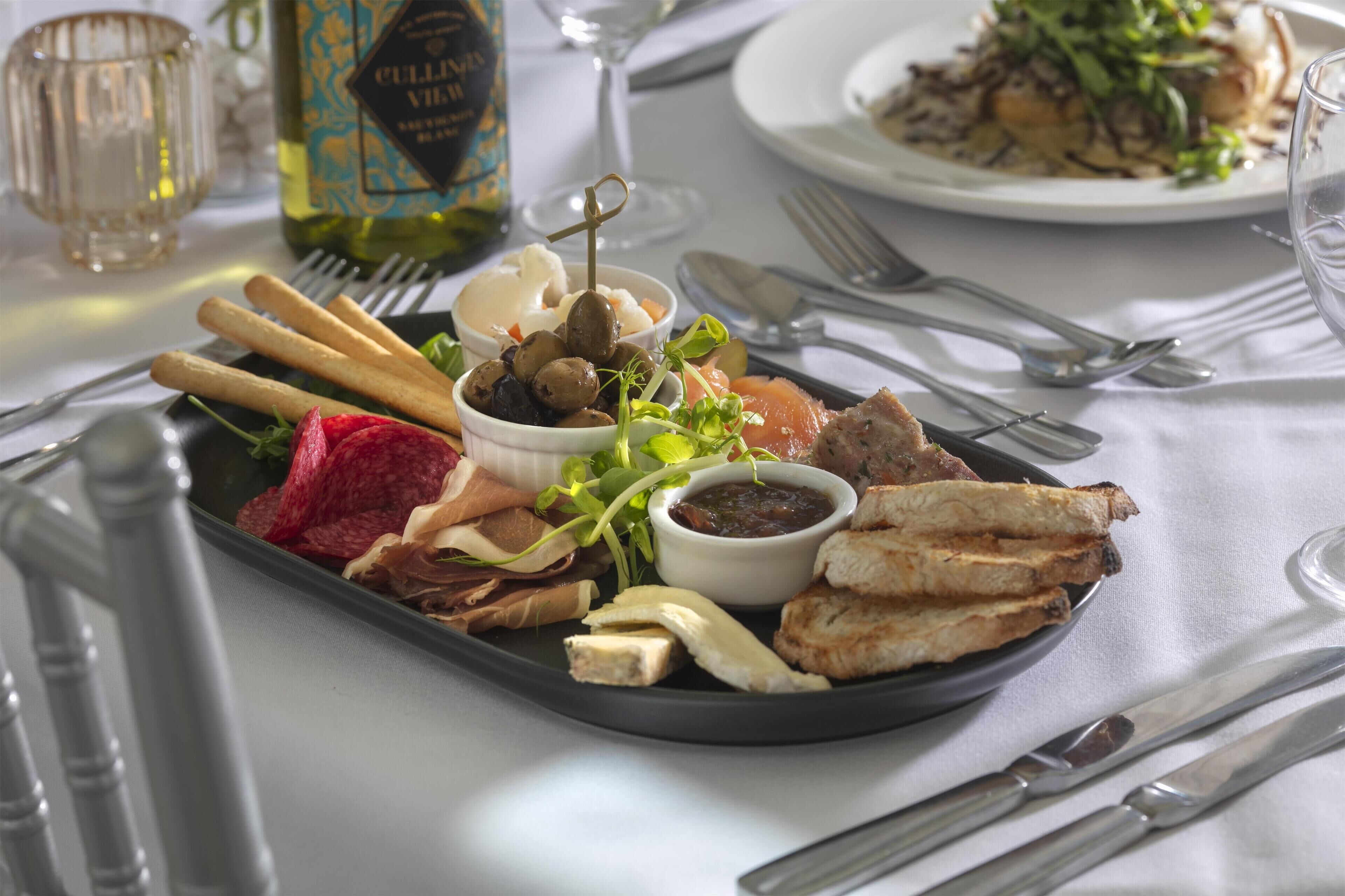 A tray of assorted food items including bread, cheese, olives, and greens, accompanied by a bottle of wine and a glass.