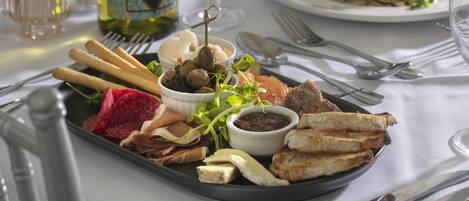 A tray of assorted food items including bread, cheese, olives, and greens, accompanied by a bottle of wine and a glass.