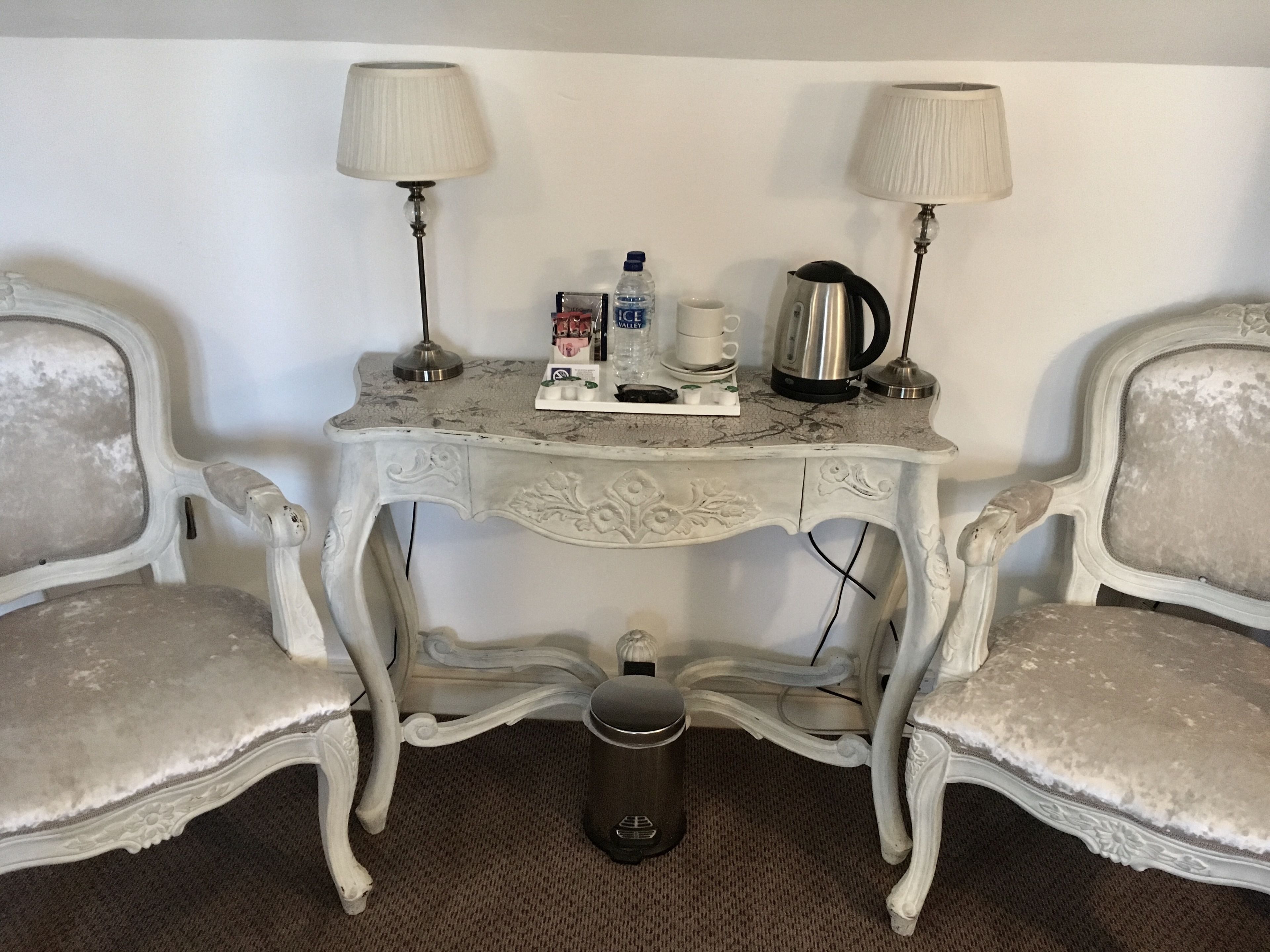 A small, white, ornate table with two lamps, a kettle, and a trash bin in a room with two matching chairs.
