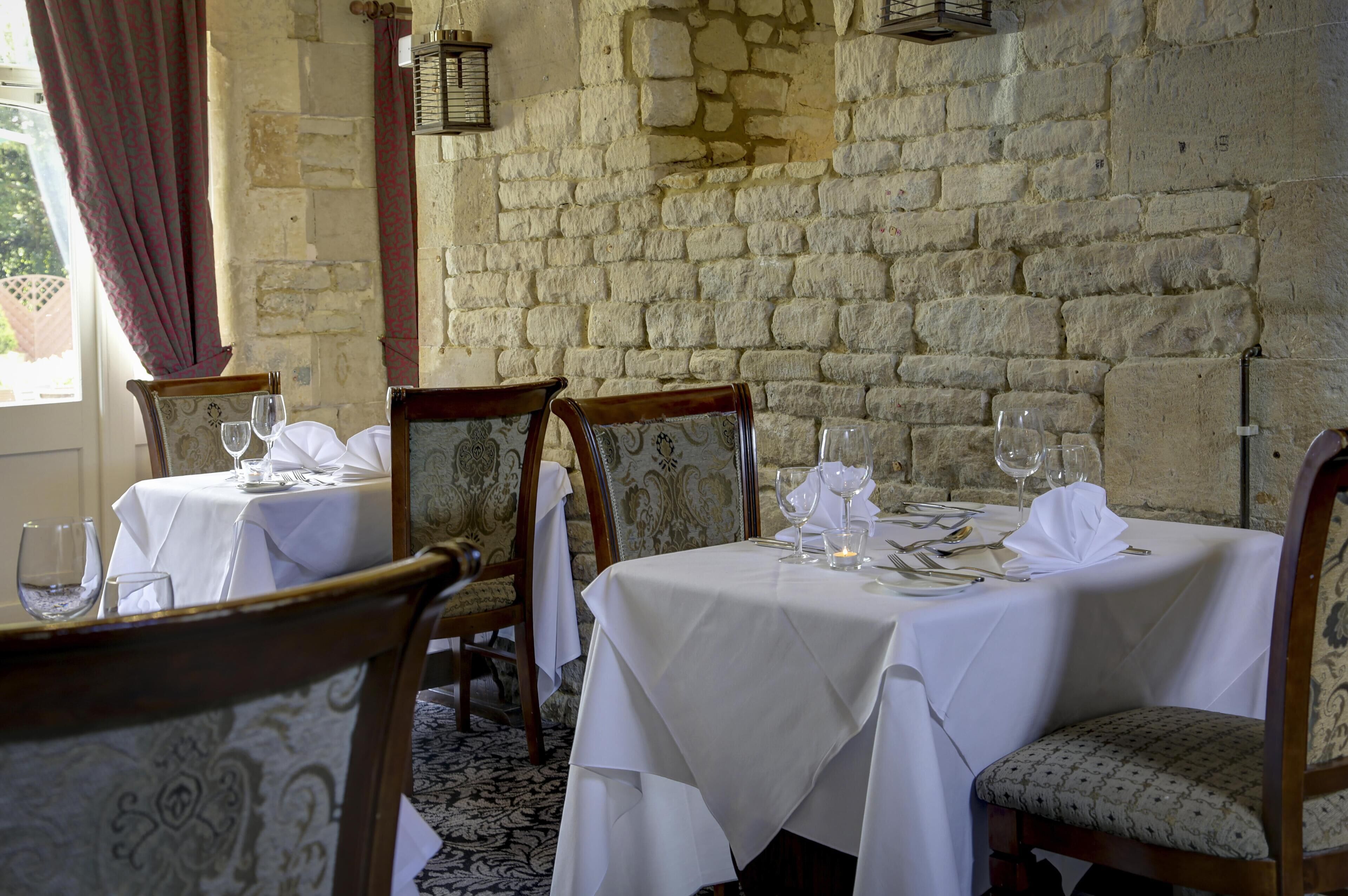 Un espace repas aux murs de pierre et aux chaises en bois, avec des tables recouvertes de nappes blanches et ornées de verres à vin.