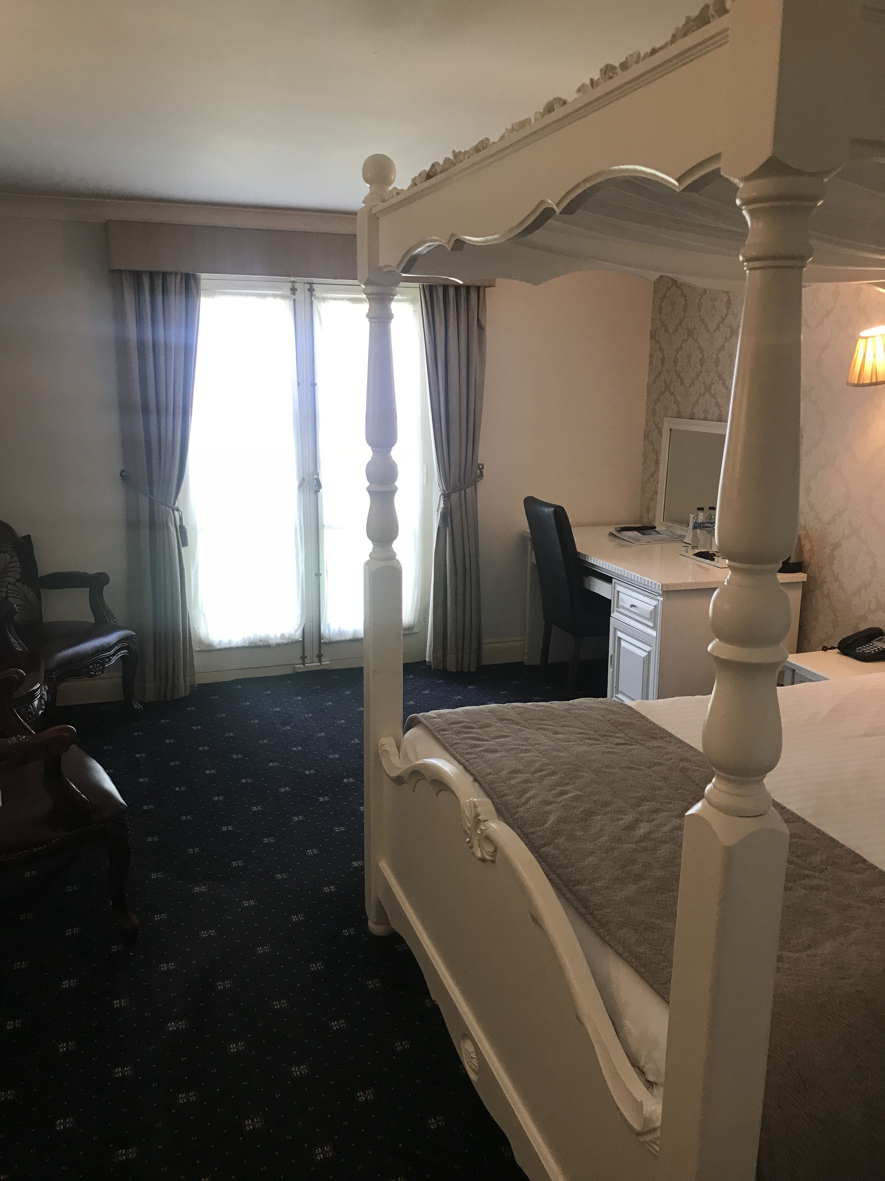 A four-poster bed with a dark patterned carpet, a desk with a chair, and a lamp.