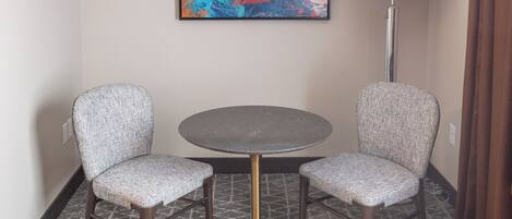 A hotel room with a round table, two patterned chairs, a floor lamp, and a large abstract painting on the wall.