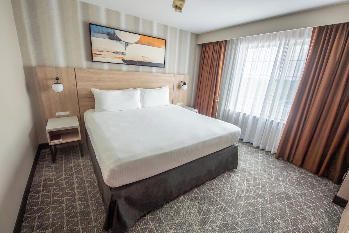 Une chambre d’hôtel avec un grand lit, une table de chevet, une lampe fixée au mur et une fenêtre avec des rideaux.