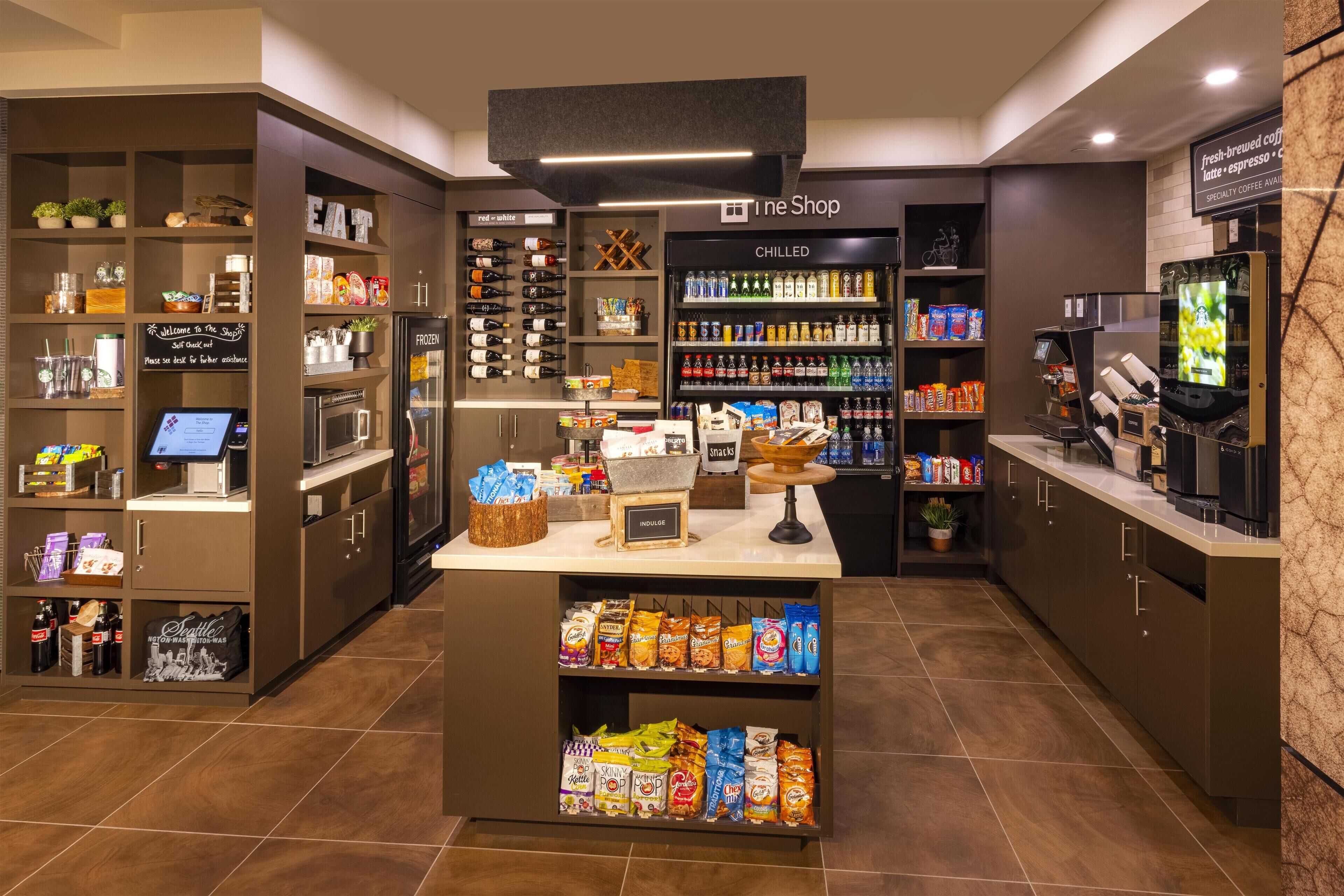 Interior de una cafetería con quiosco de autoservicio, sección refrigerada y estanterías de aperitivos.