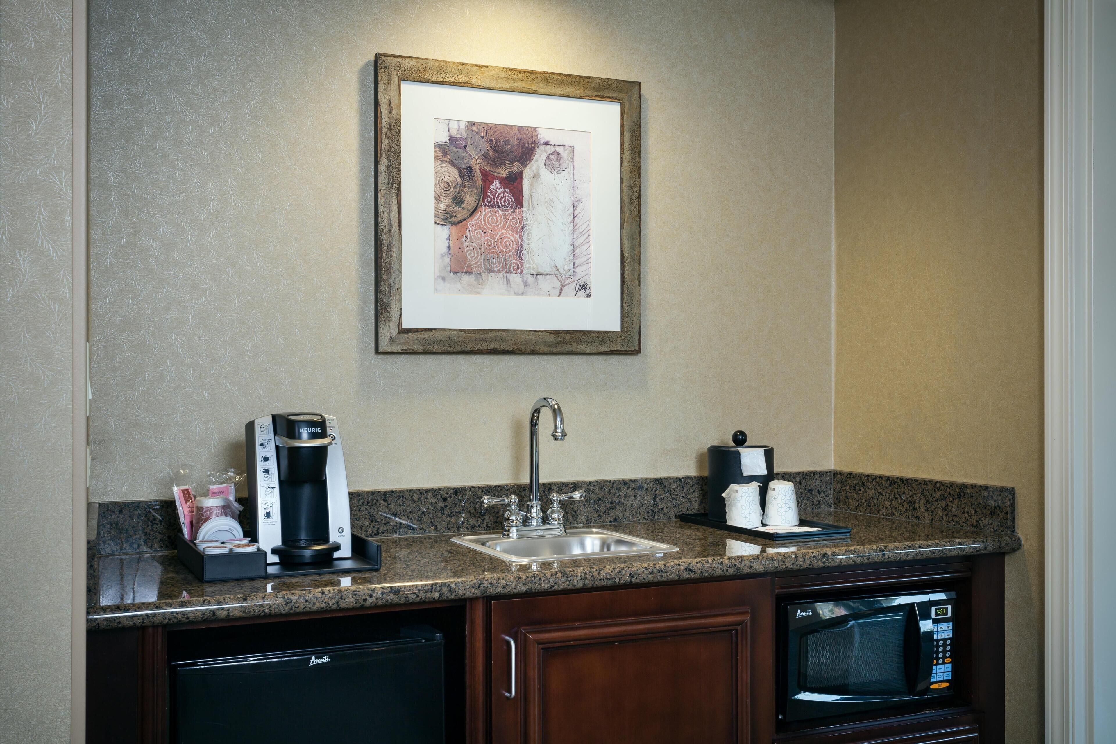 A kitchen with a coffee maker, sink, microwave, and a framed artwork on the wall.
