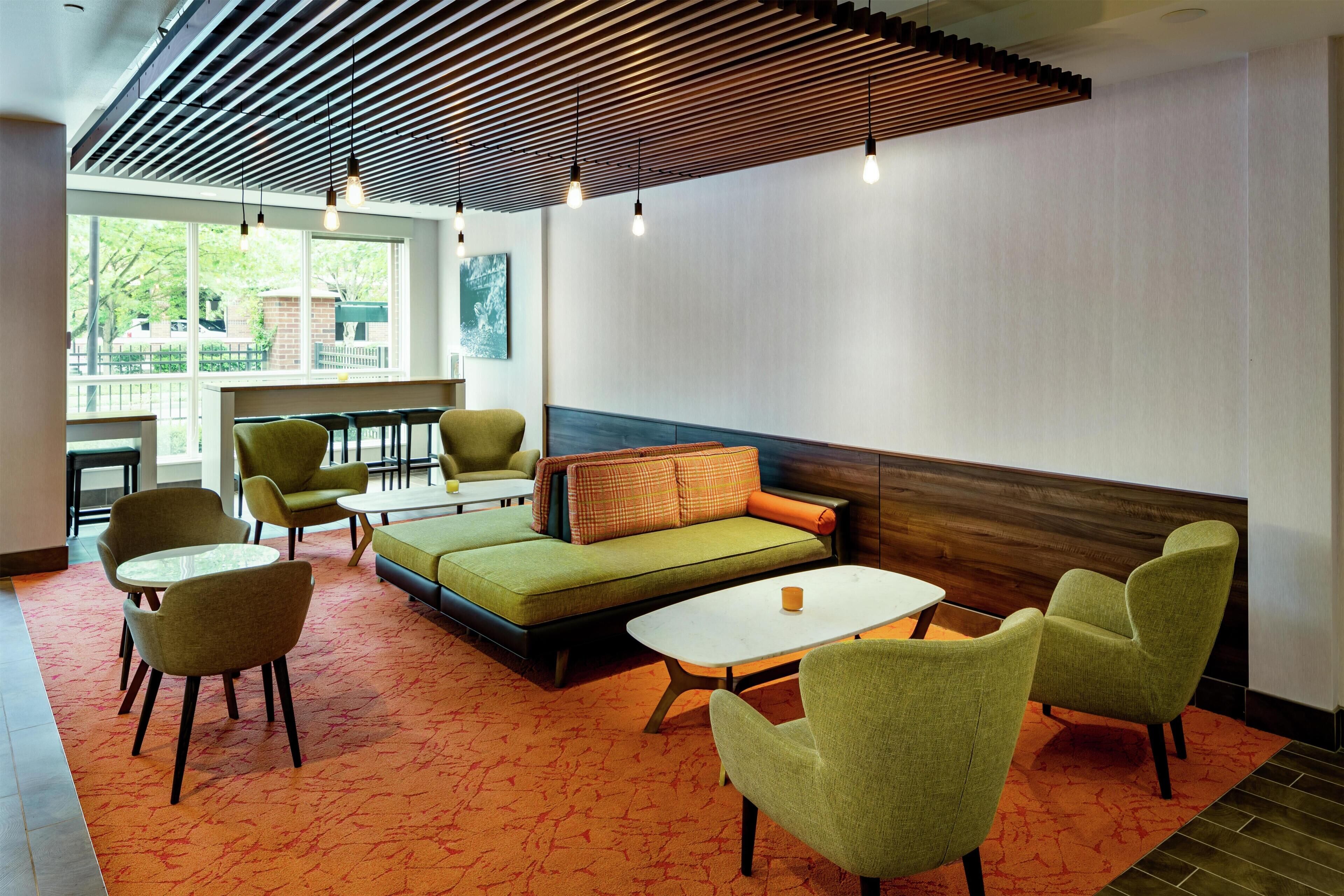 A modern lounge area with a sofa, chairs, and tables, featuring a wooden ceiling and large windows.