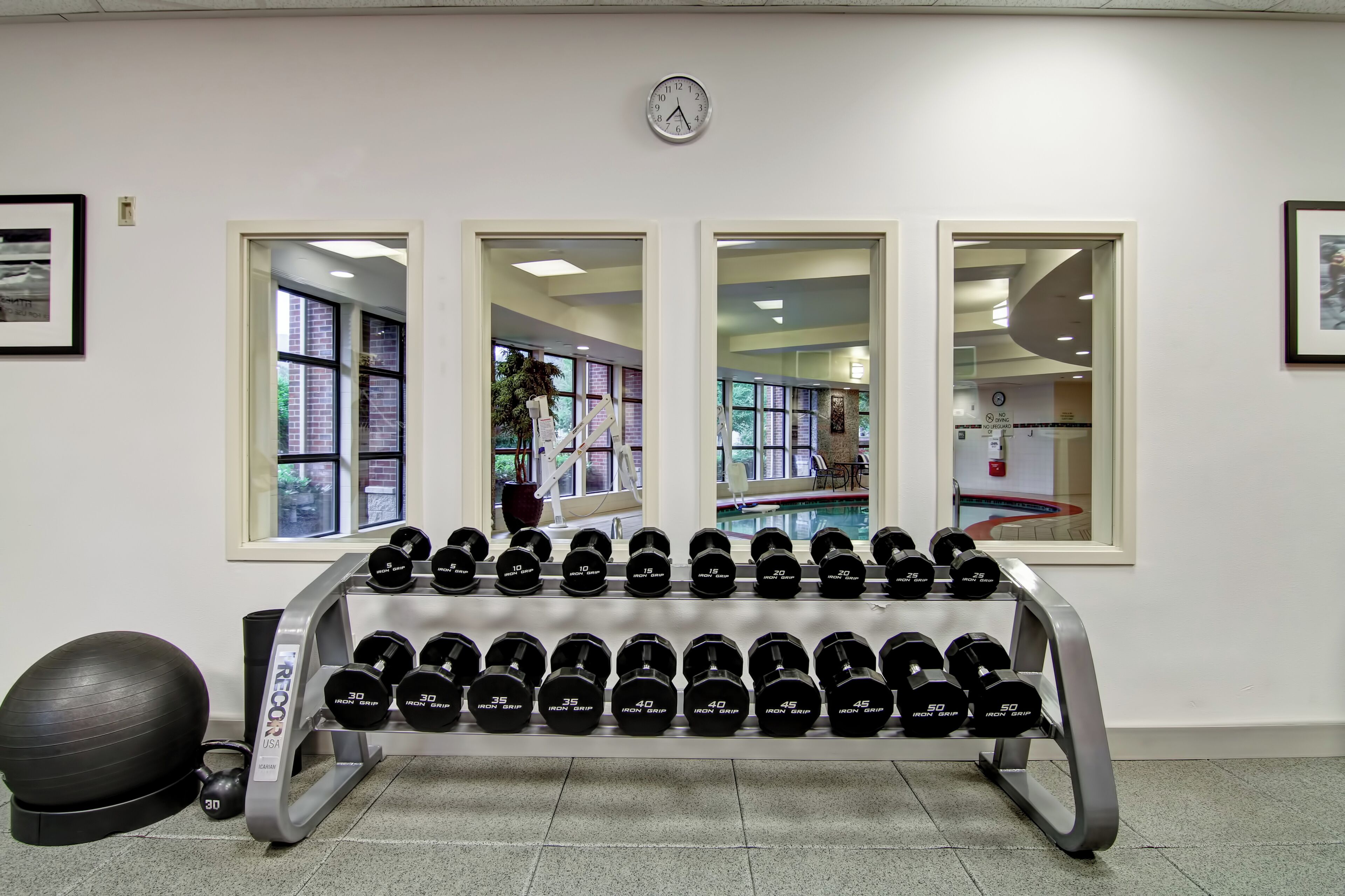 A gym with a wall-mounted clock, large windows, and a rack of dumbbells.