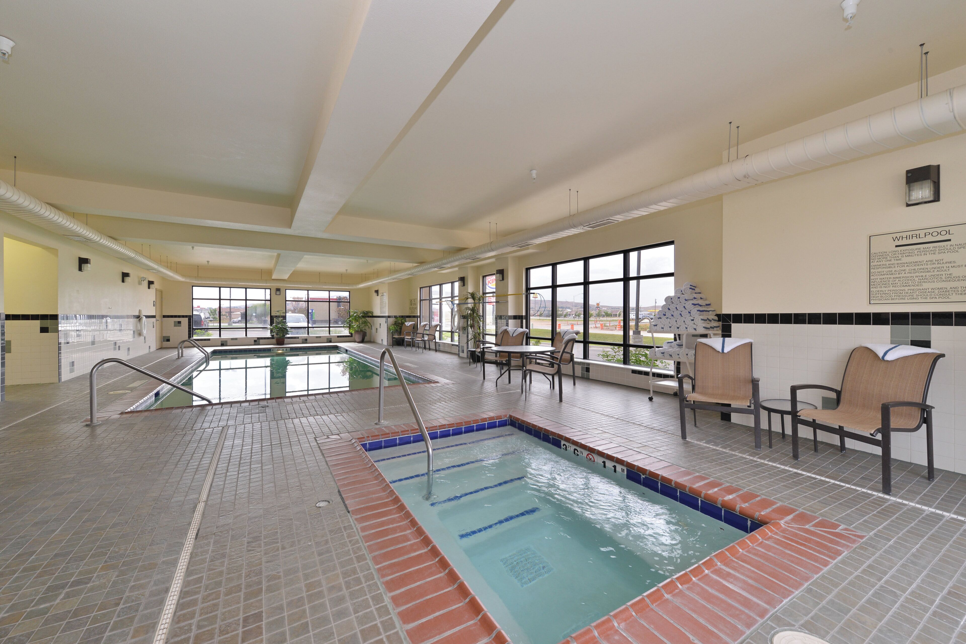 An indoor swimming pool with a tiled floor, a pool ladder, and a seating area with chairs.