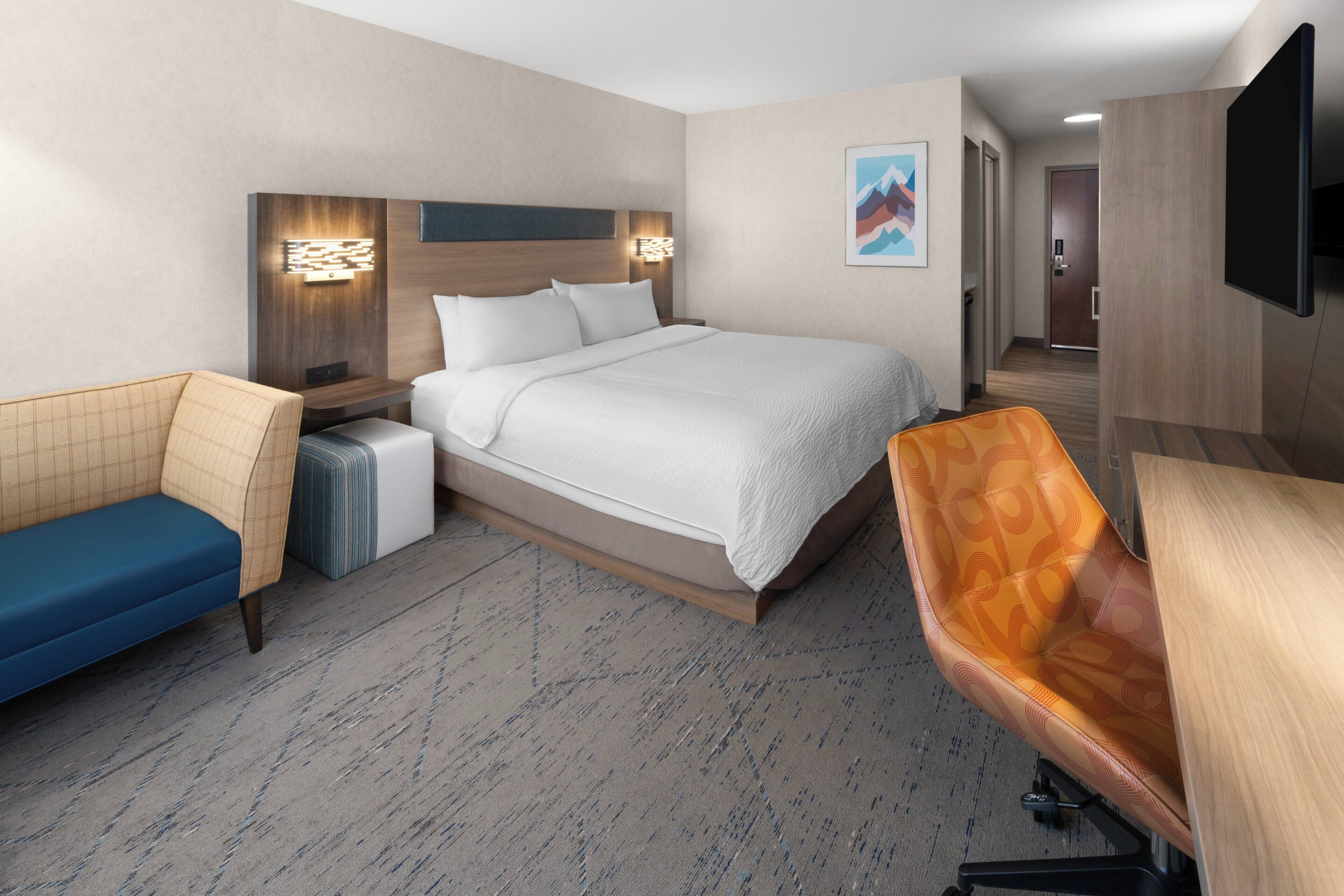 A hotel room with a bed, a sofa, a chair, a TV, and a wall-mounted light fixture.
