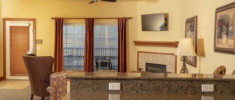 A hotel room with a large window, a flat-screen TV, a fireplace, and a seating area with a chair and a table.