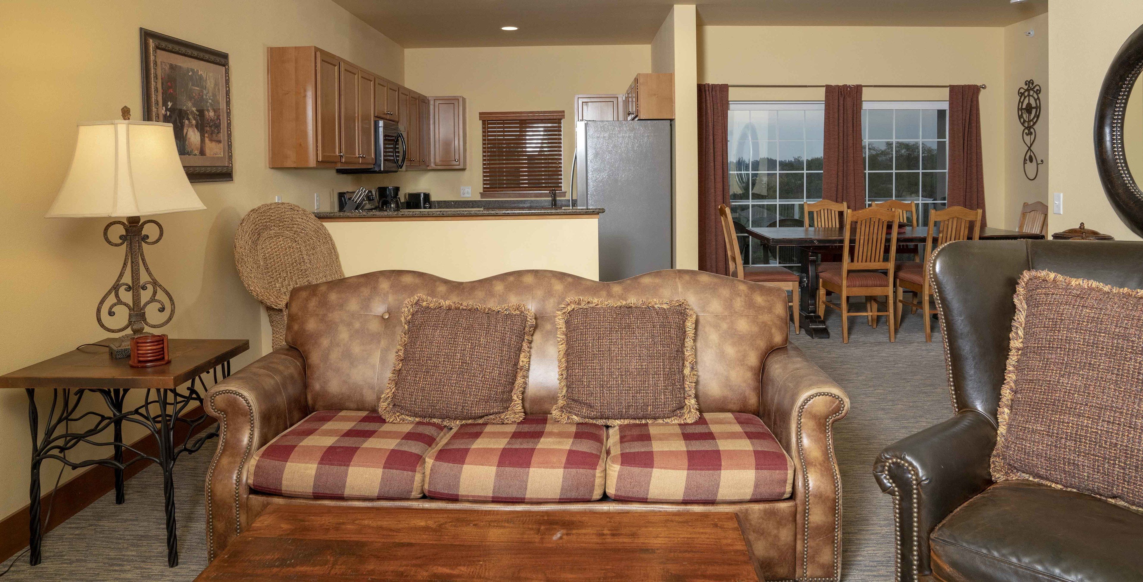 A living room with a sofa, a coffee table, a lamp, a dining area with a table and chairs, and a kitchen with cabinets and appliances.
