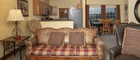 A living room with a sofa, a coffee table, a lamp, a dining area with a table and chairs, and a kitchen with cabinets and appliances.