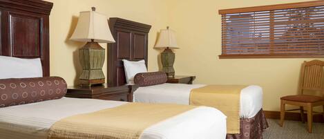 A hotel room with two single beds, each with a wooden headboard, a small table, two lamps, and a window with blinds.