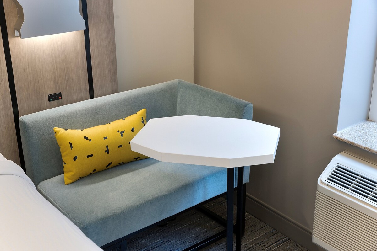 A room with a grey sofa, a yellow pillow, a white table, and a white air conditioning unit.