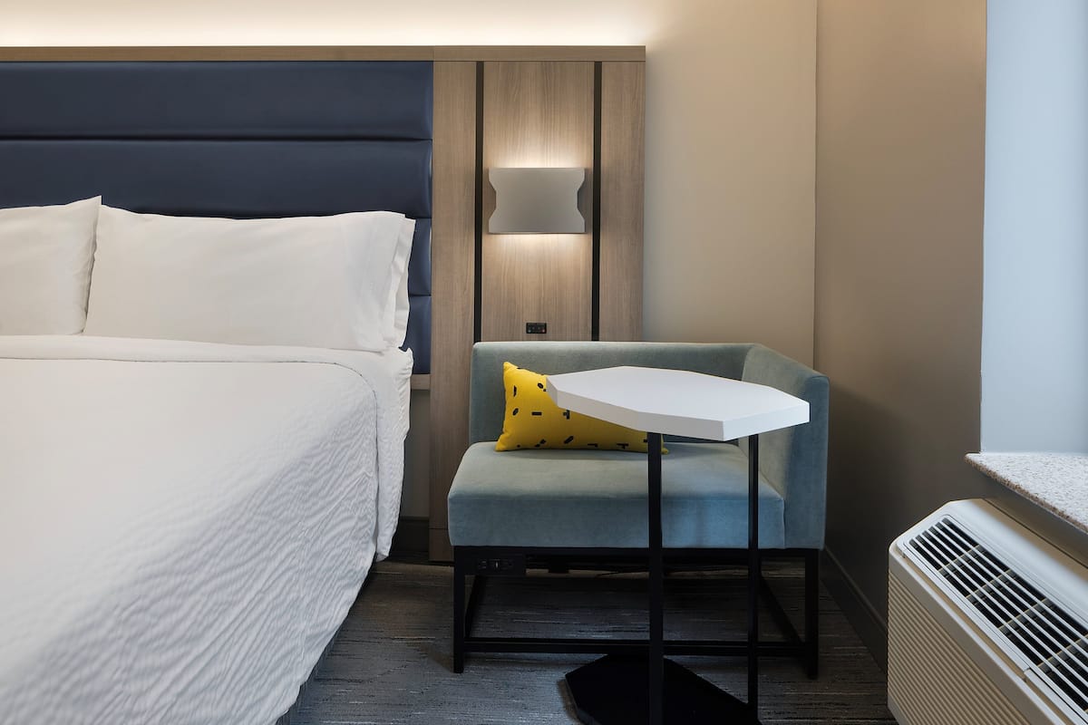 A hotel room with a bed, a small table, a chair, a lamp, and a heater.