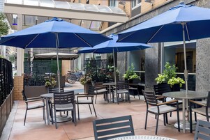 Un patio al aire libre con mesas y sillas bajo sombrillas azules, rodeado de plantas en macetas y un muro de ladrillo.