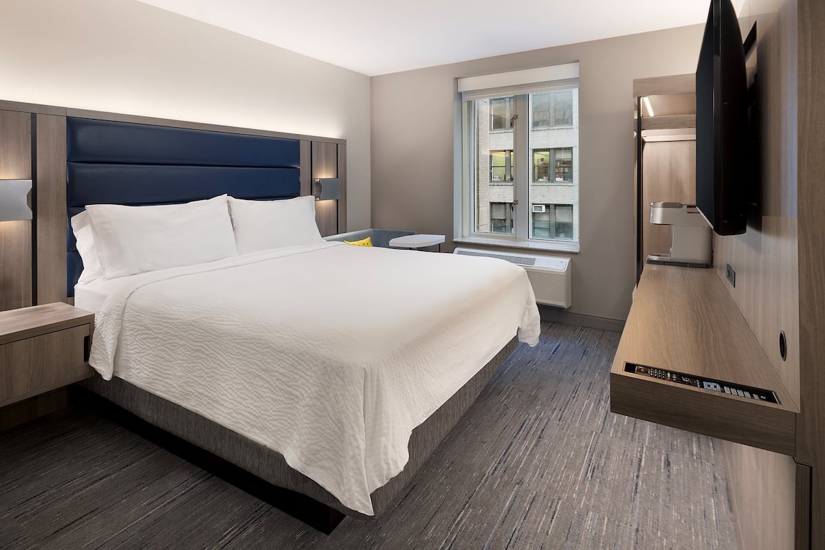 A modern hotel room with a large bed, a flat-screen TV mounted on the wall, and a window with a view of the city.