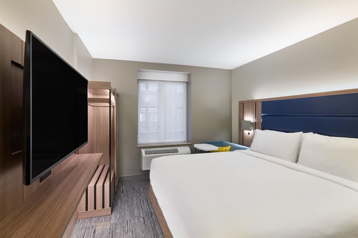 A hotel room with a large bed, a flat-screen TV, a small table, and a window with blinds.