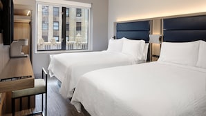 Une chambre d’hôtel avec deux lits, un bureau et une grande fenêtre donnant sur un immeuble.