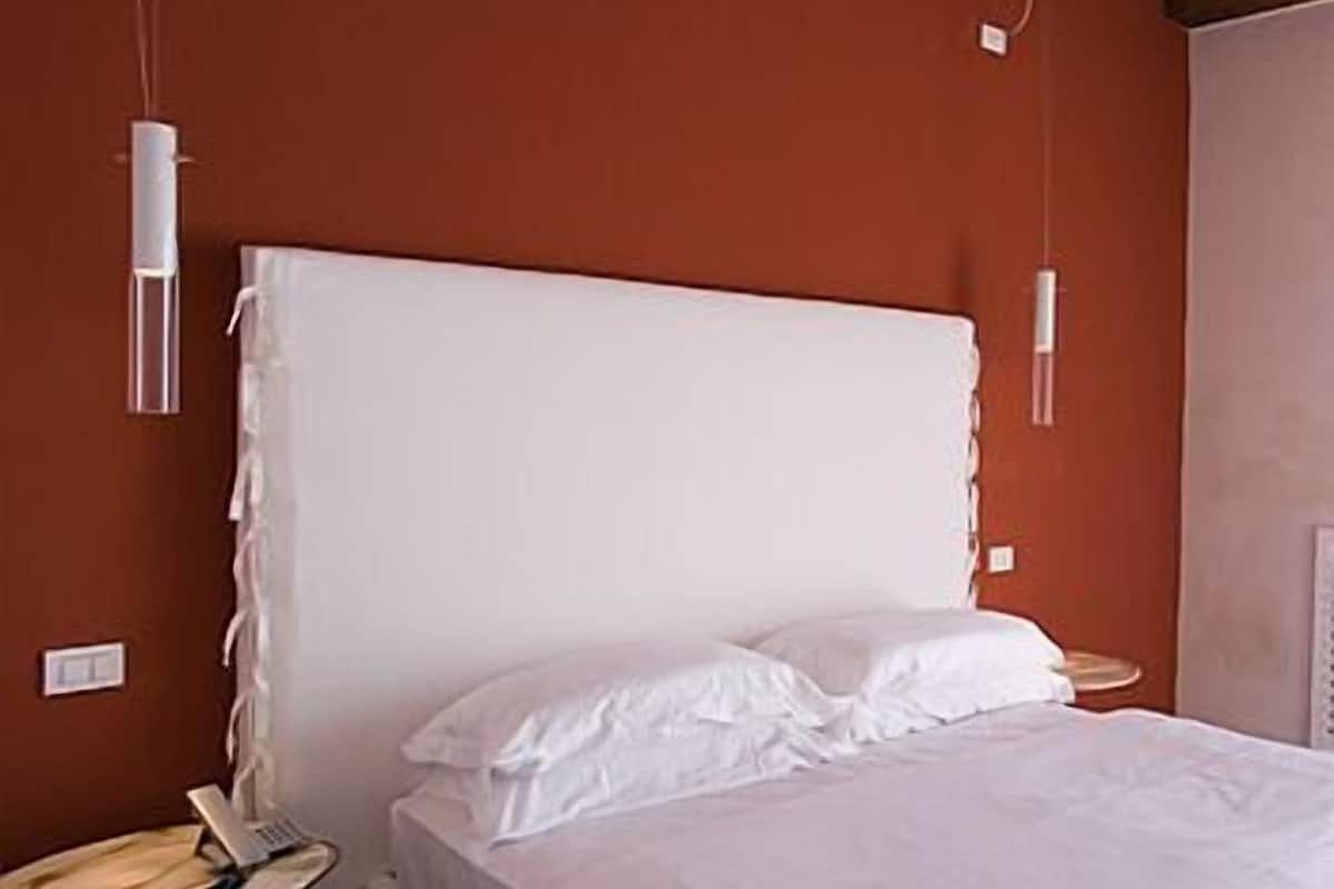 A bedroom with a large bed, white bedding, a bedside table with a lamp, and a wall socket.