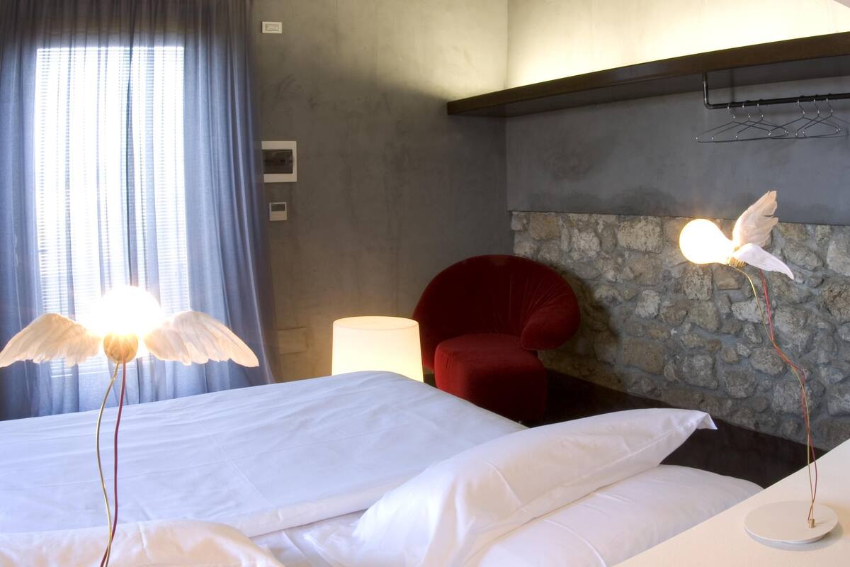 A hotel room with a bed, a red chair, a stone wall, and a decorative light fixture.