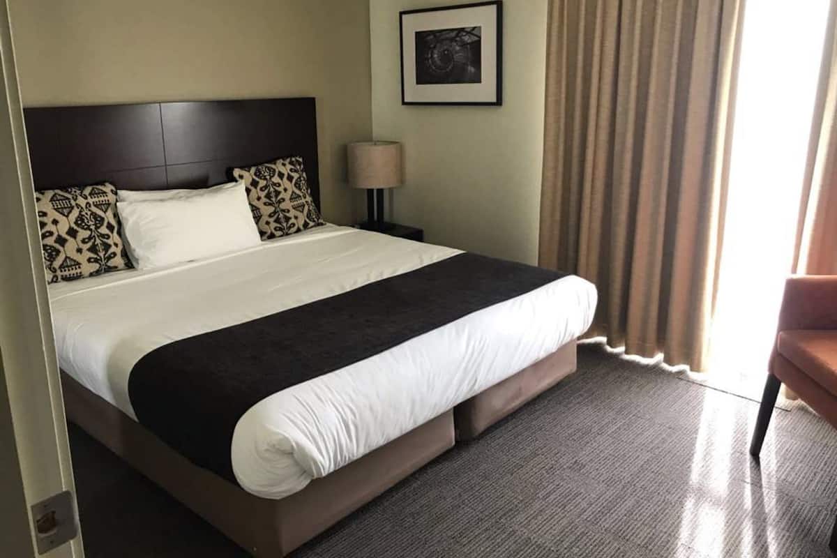 Une chambre d’hôtel avec un grand lit, une table de chevet avec une lampe, un tableau encadré au mur et une fenêtre avec des rideaux.
