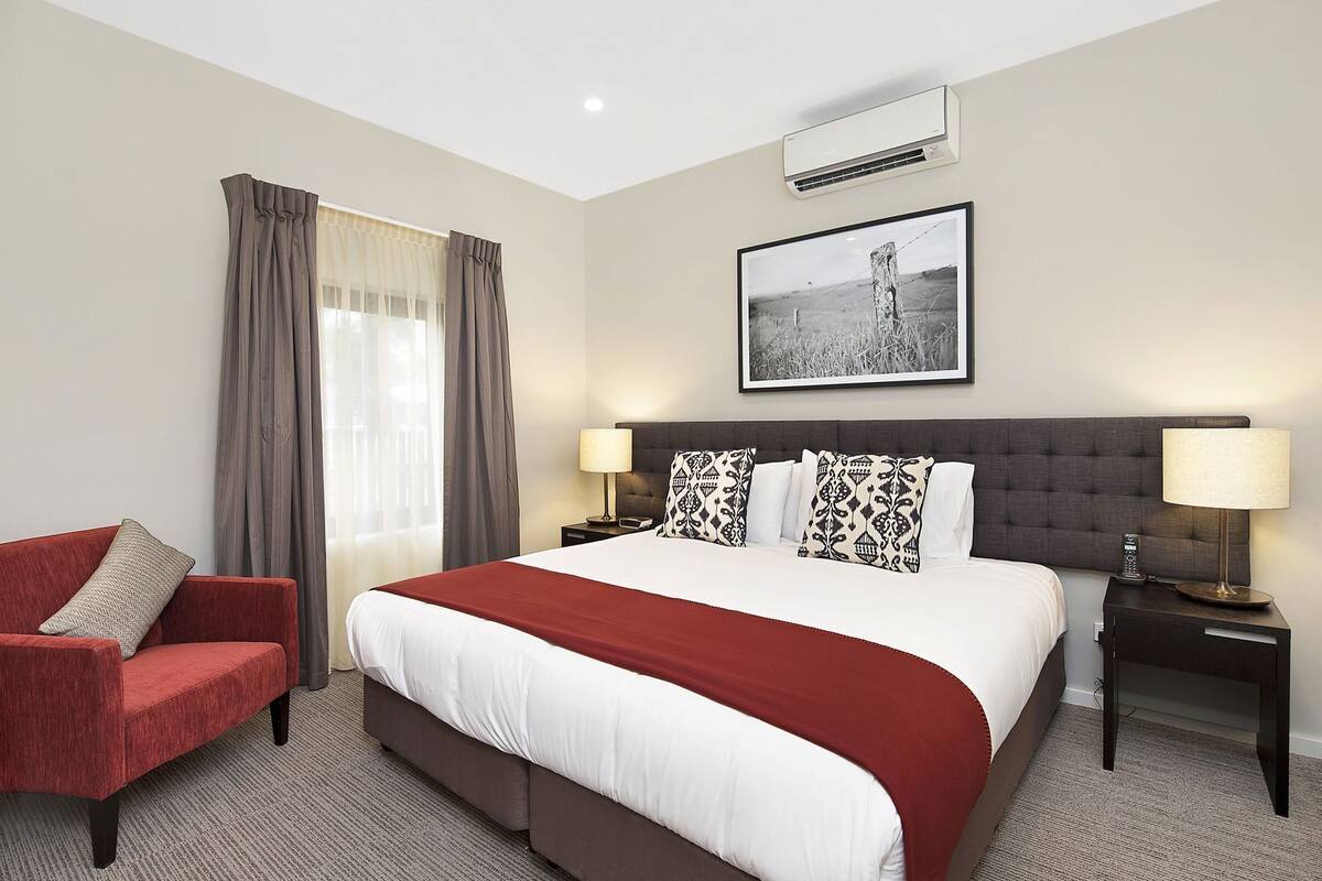 Une chambre d’hôtel avec un grand lit, un fauteuil rouge, une table de chevet avec une lampe et un tableau encadré au mur.
