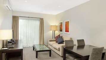 A hotel room with a sofa, a small table, a larger dining table, and a lamp.