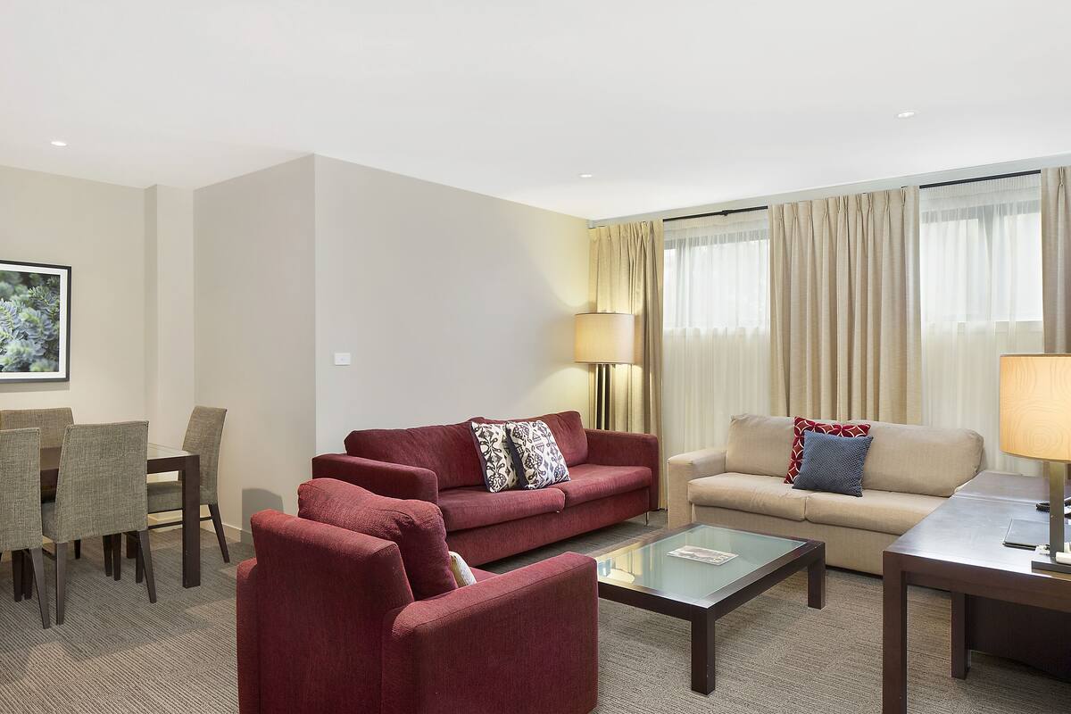 Une chambre d’hôtel avec un canapé rouge, un canapé beige et une table basse en verre. On y trouve également une table à manger avec des chaises et une lampe.
