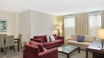 A hotel room with a red sofa, beige sofa, and a glass coffee table. There is a dining table with chairs and a lamp.