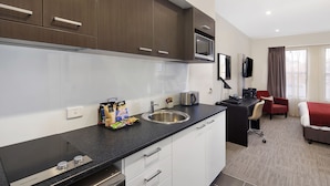 Cocina moderna con microondas integrado, un fregadero y una cafetera. Hay una pequeña zona de estar con una silla y una cama al fondo.