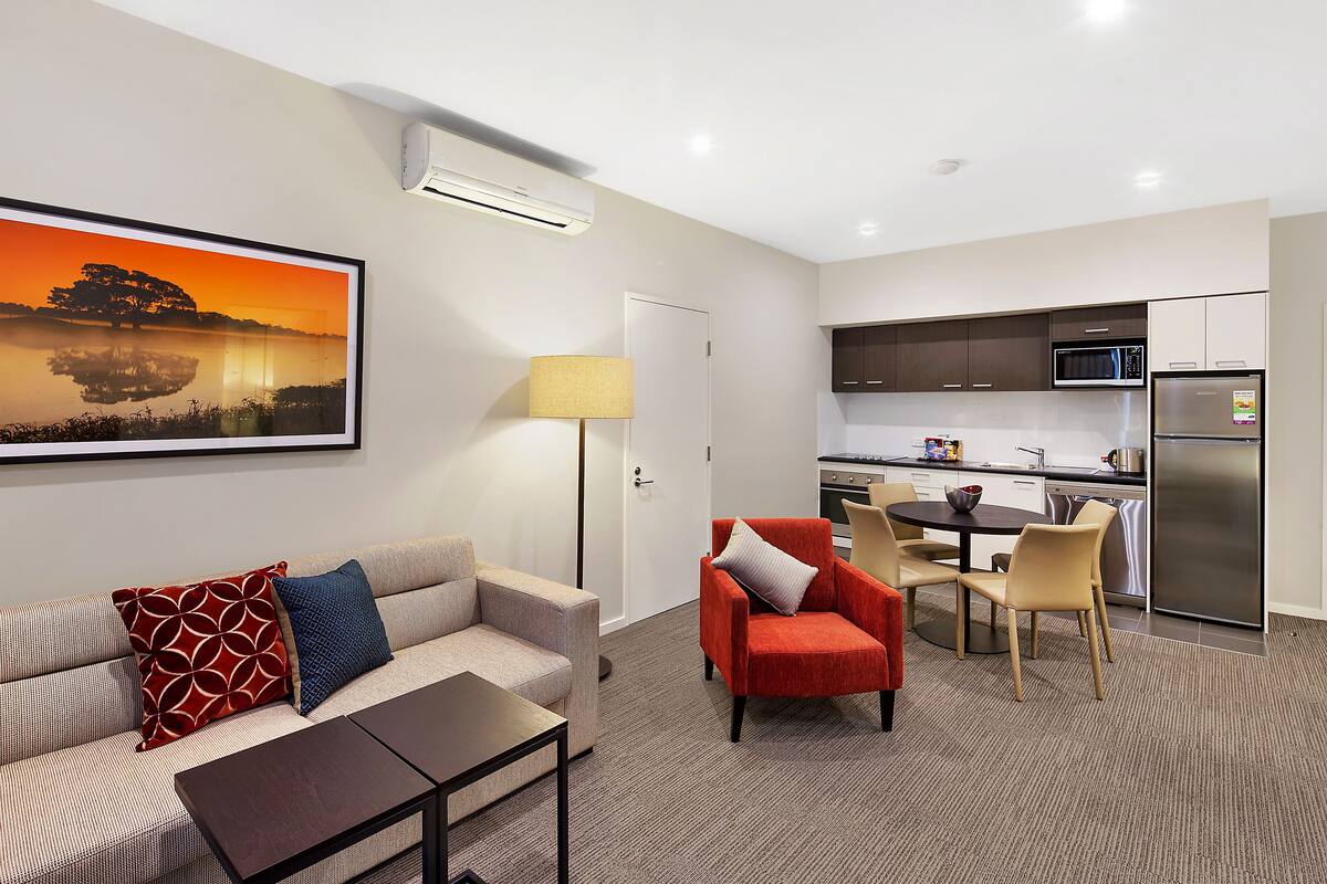 Une chambre d’hôtel moderne équipée d’un canapé, d’un fauteuil, d’une table à manger et d’un coin cuisine comprenant un réfrigérateur et un four à micro-ondes.