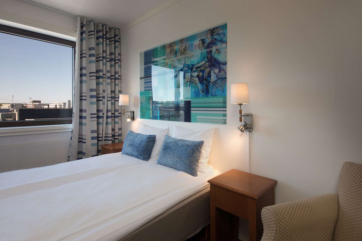 A hotel room with a large bed, two pillows, a nightstand, a lamp, a chair, and a painting on the wall.