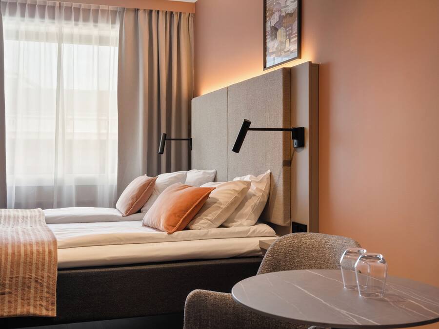 A hotel room with a bed, a table with chairs, a headboard, a wall-mounted lamp, and a window with curtains.