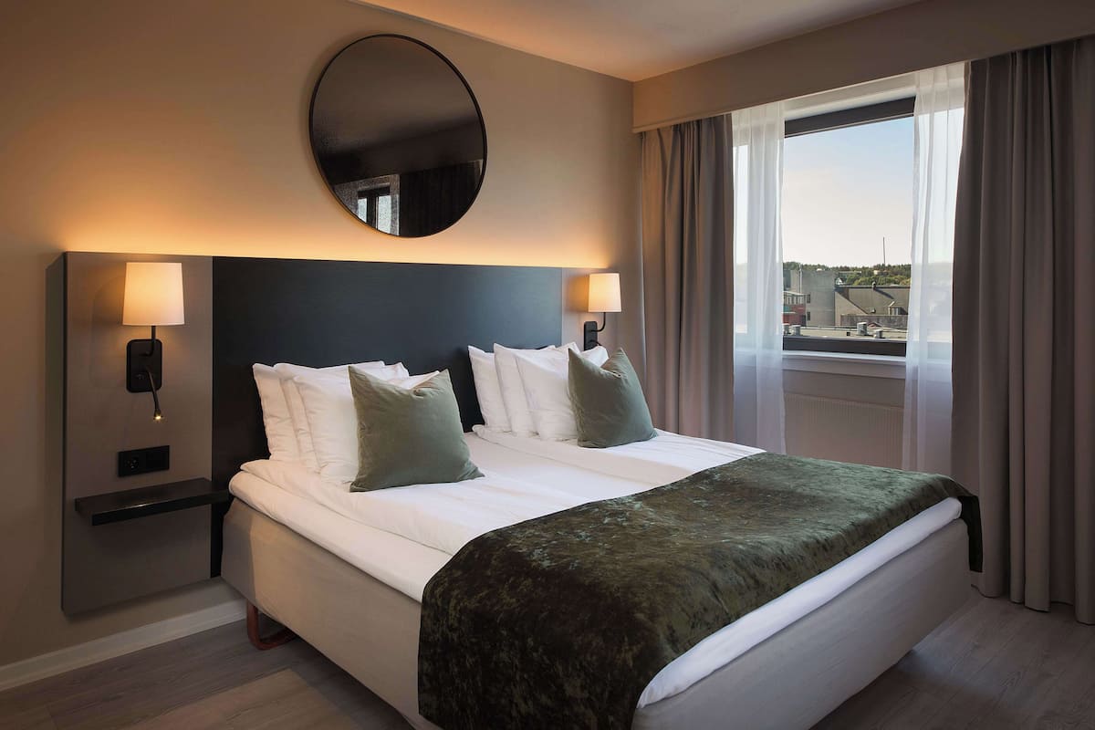 A hotel room with a large bed, two bedside lamps, a round mirror, and a view of the city through the window.