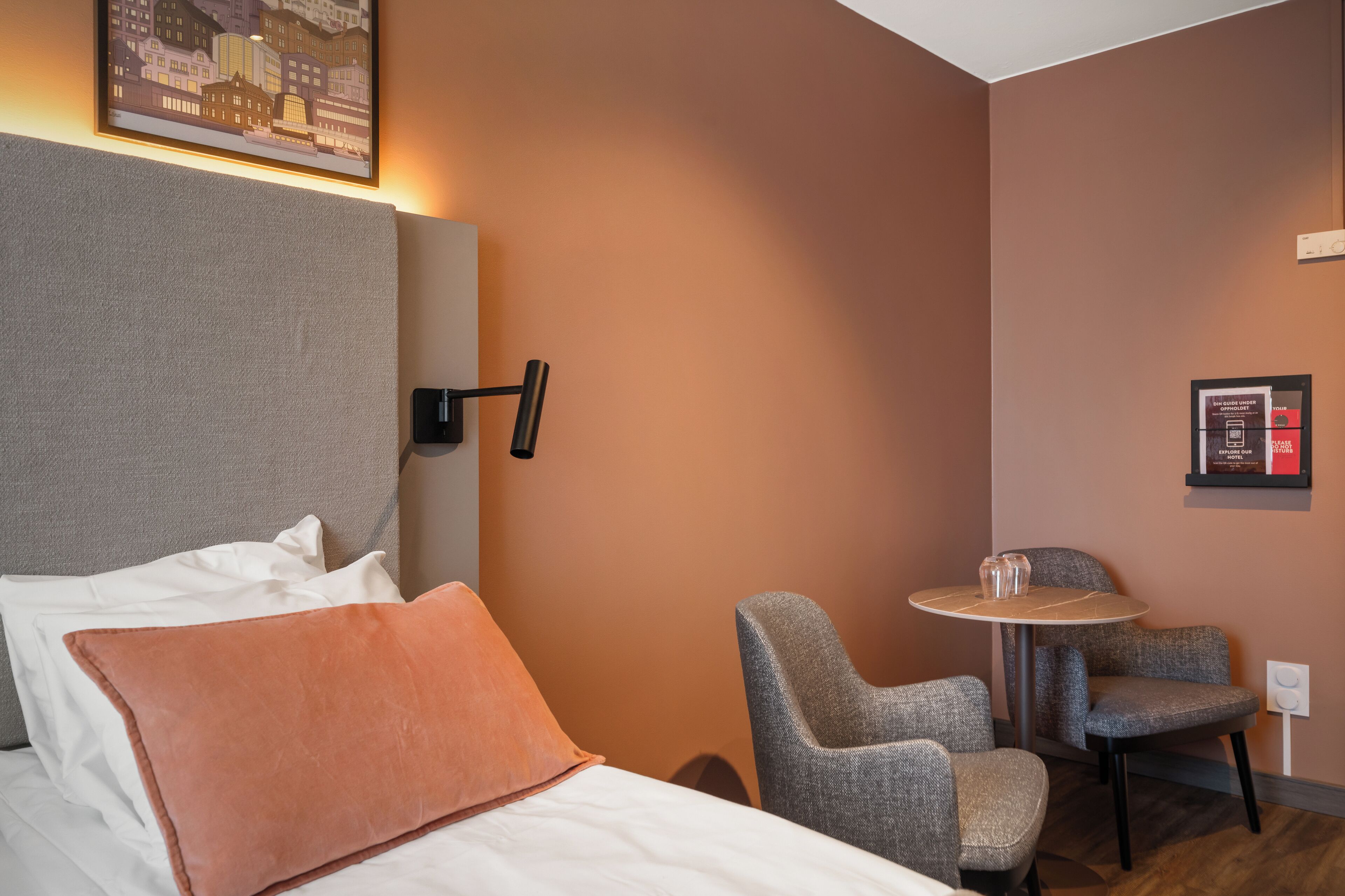 A hotel room with a bed, a gray armchair, a small round table, and a wall-mounted light fixture.