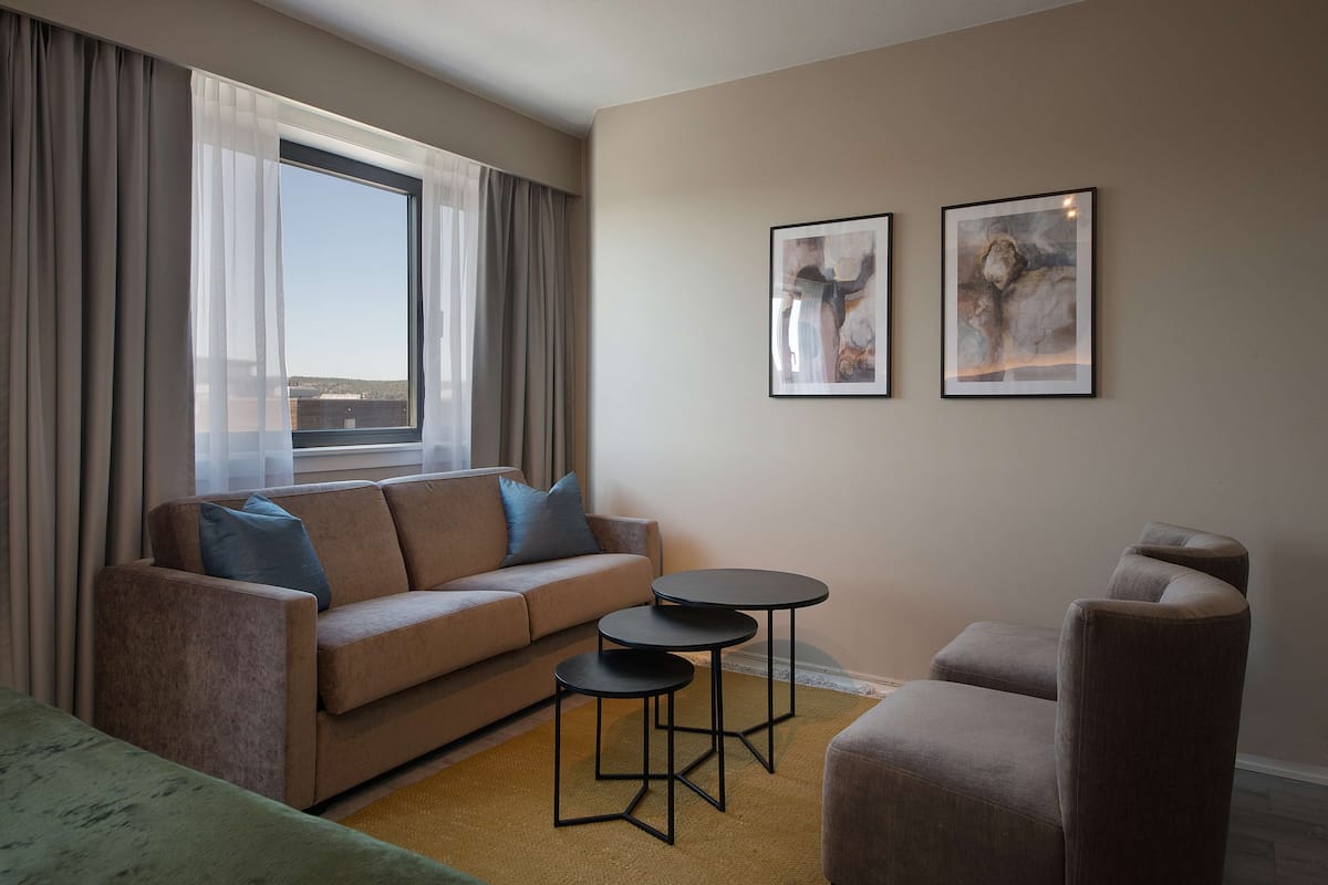 A modern living room with a sofa, two small tables, and a chair. There are two framed artworks on the wall and a large window with curtains.