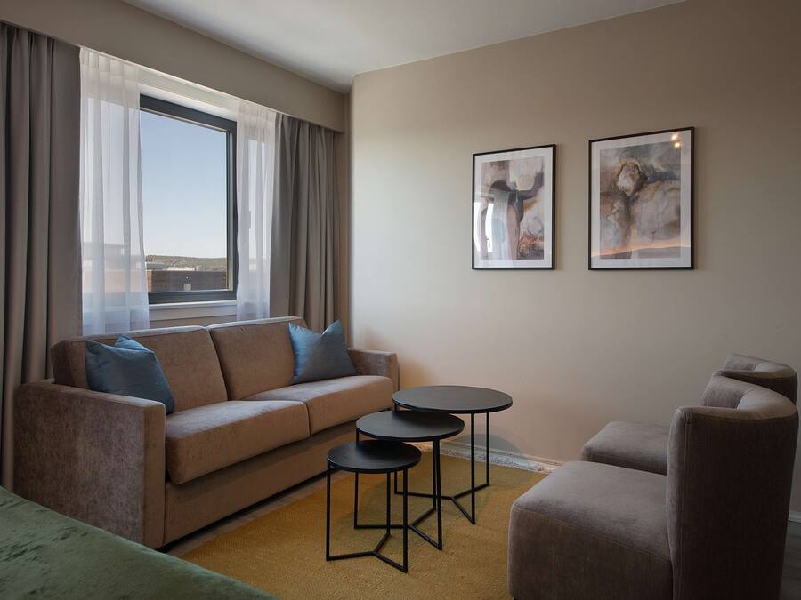 A modern living room with a sofa, two small tables, and a chair. There are two framed artworks on the wall and a large window with curtains.