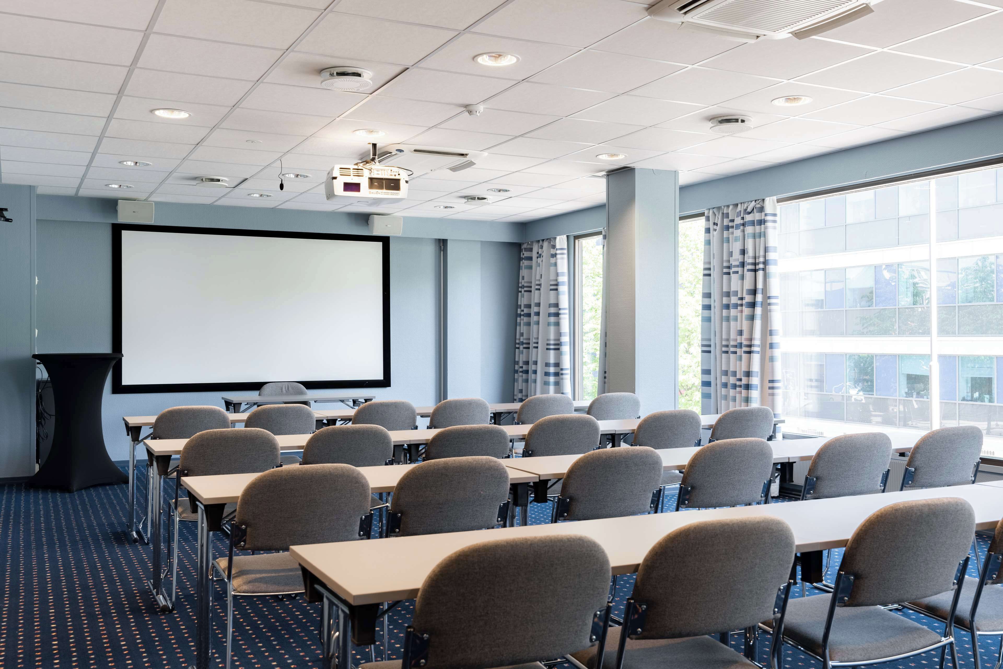 Un salón de conferencias con filas de mesas y sillas, una pantalla de proyector y ventanas amplias.