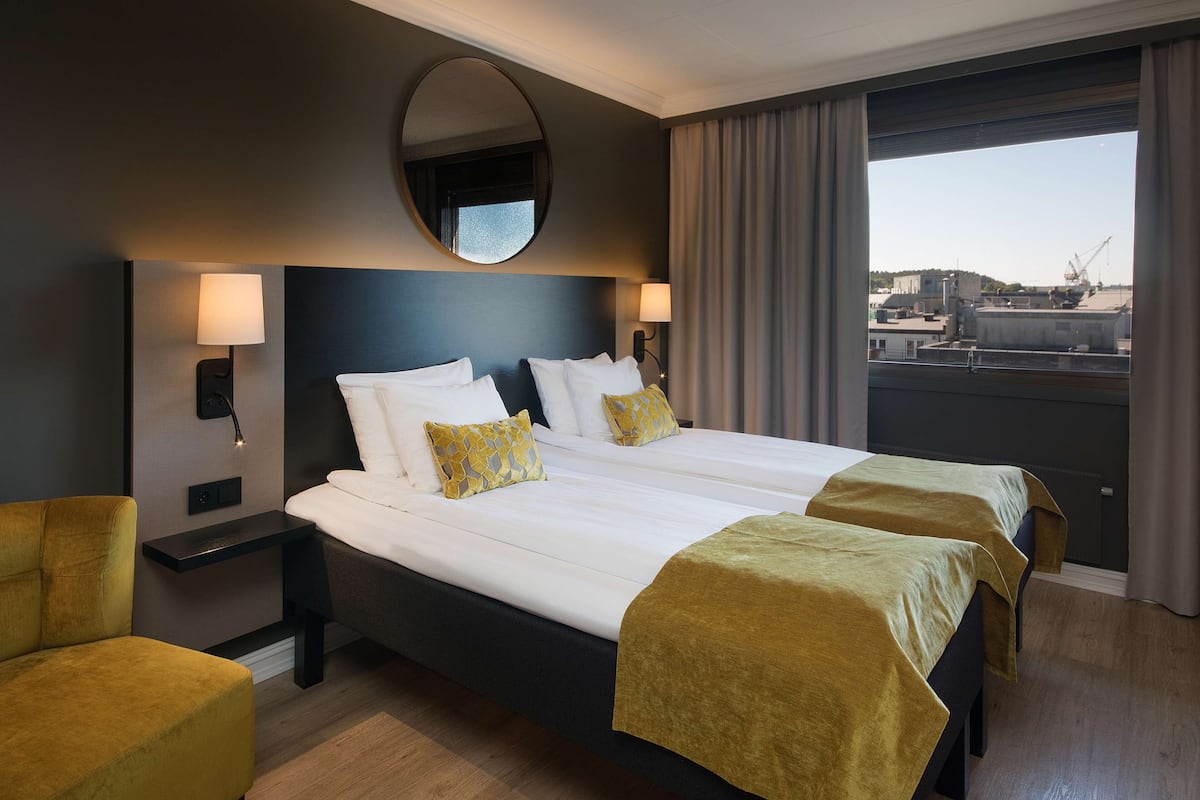 A hotel room with a large bed, two bedside tables, a round mirror, and a view of the city through the window.