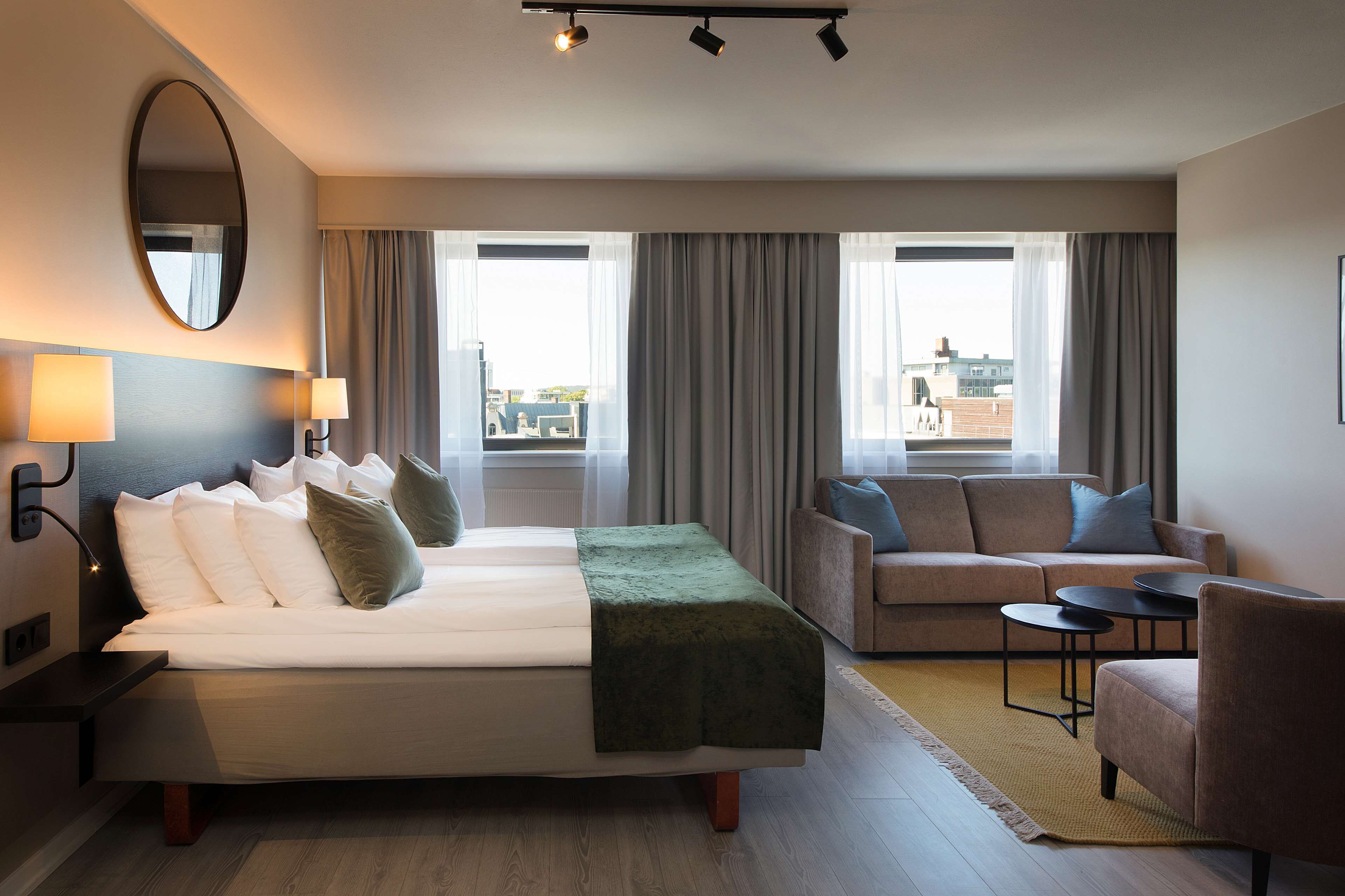 Habitación de hotel con cama, sofá, sillón y una pequeña mesa. Hay dos ventanas con cortinas, un espejo en la pared y una lámpara en la mesita de noche.