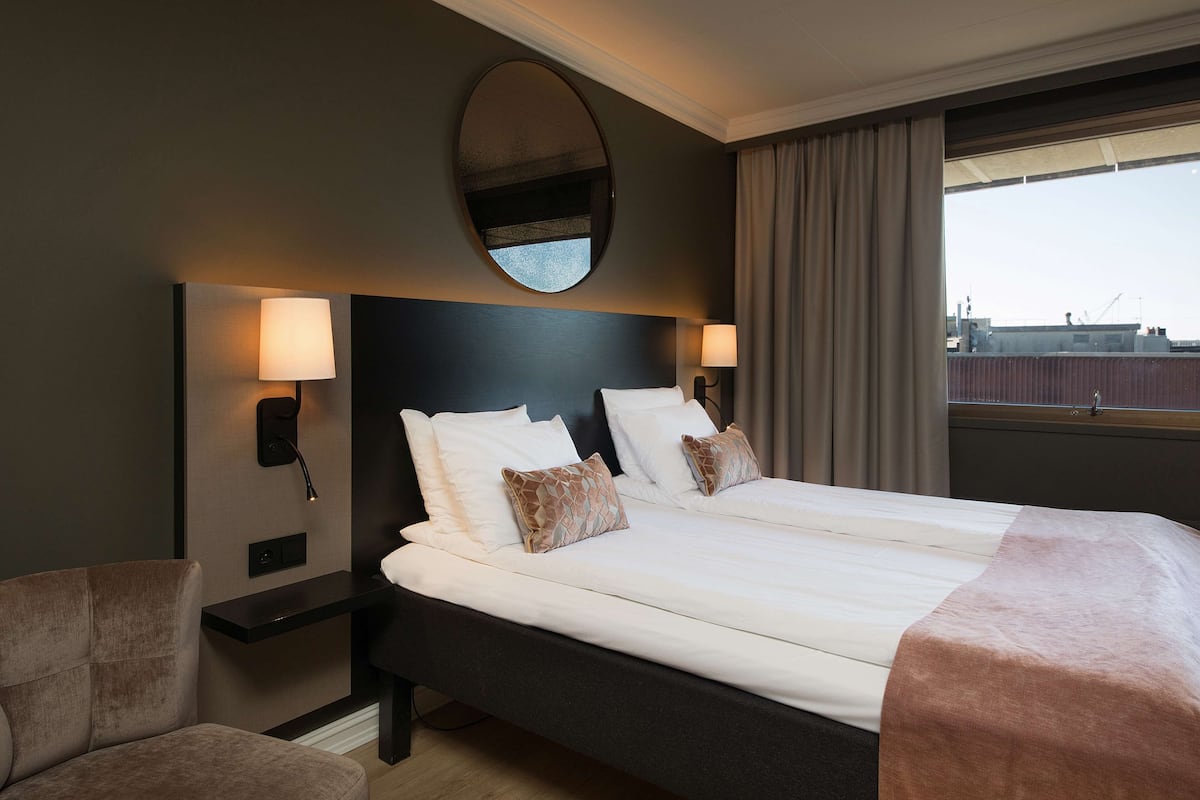 A hotel room with a large bed, two bedside tables with lamps, a round mirror above the bed, and a view of the city through the window.