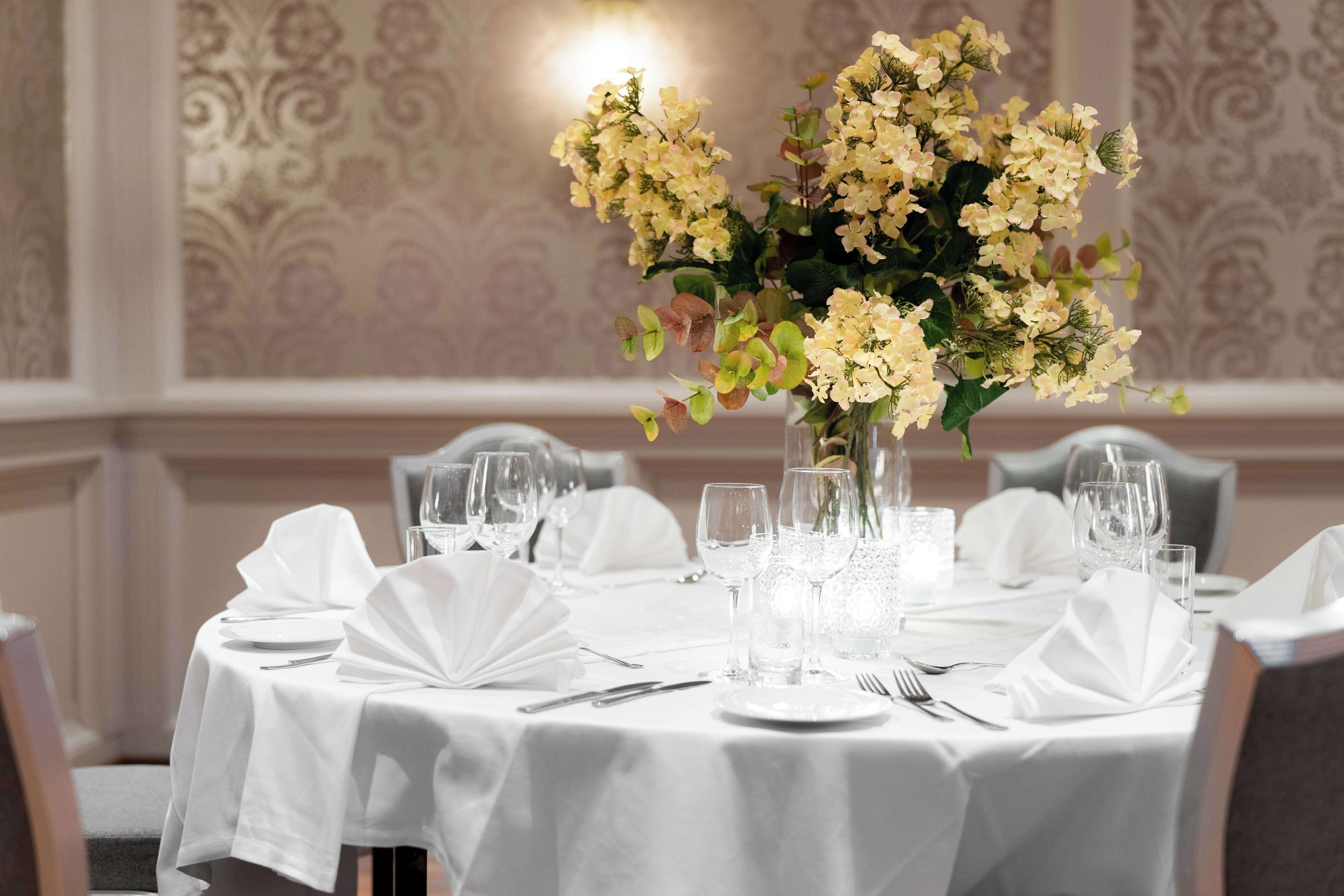 Una mesa de comedor dispuesta para una comida formal, con manteles blancos, cristalería y un centro de mesa con flores amarillas.