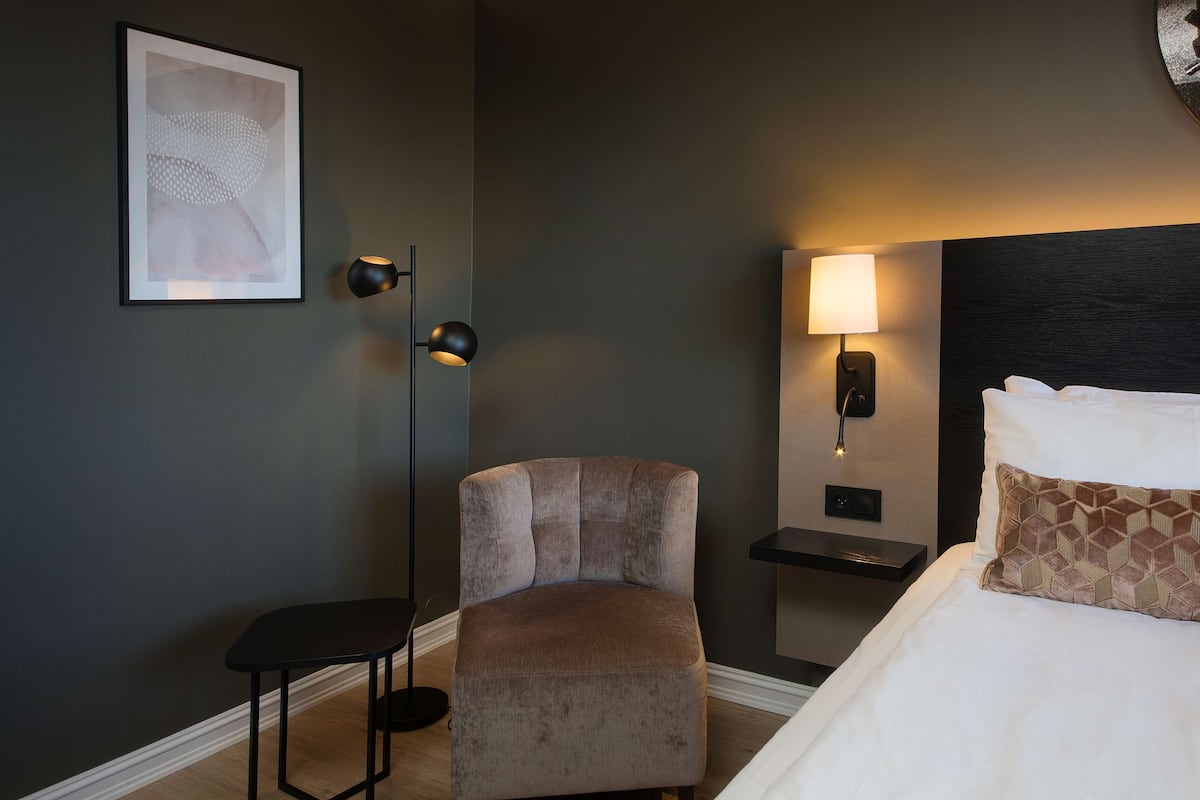 A hotel room with a bed, a chair, a nightstand, a wall-mounted lamp, and a framed artwork.